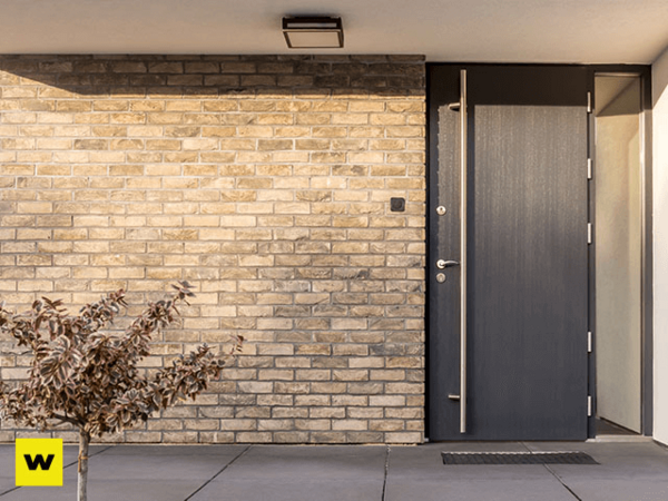 Moderne Haustür aus dunklem Material vor heller Klinkerfassade mit kleinem Baum davor.