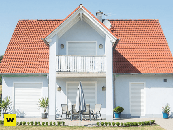 Modernes Einfamilienhaus mit rotem Dach, Terrasse und Rollläden bei Sonnenschein.